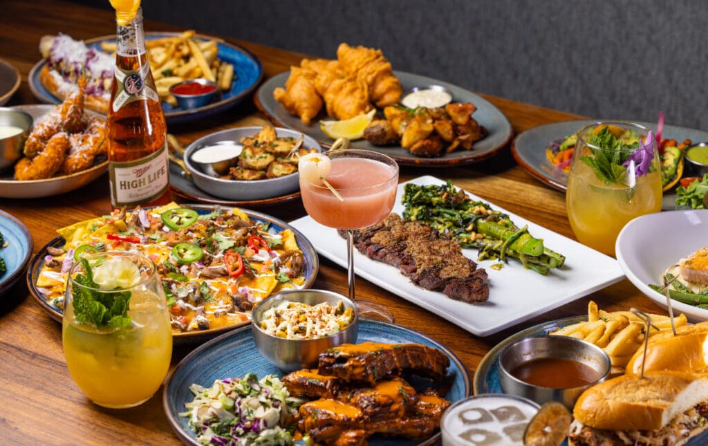 A table at Eureka! in Novus Place Tempe with pizza, steaks, cocktails and other plates of food.