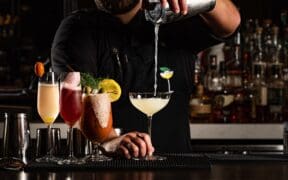 A row of cocktail glasses sits on a bar top while a bartender pours a drink in a martini glass.