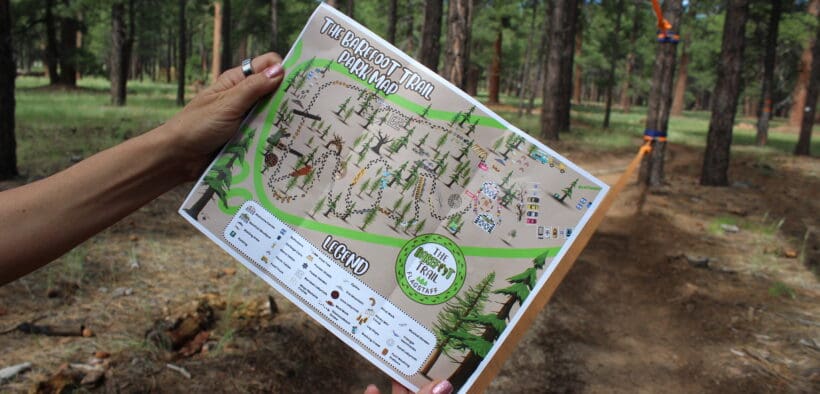 A person is holding up a map of the Flagstaff Barefoot Trail, with forest trees in the background.