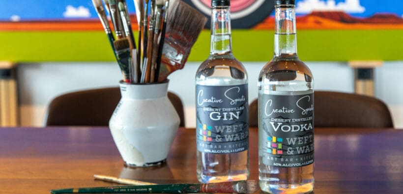 Creative Spirits liquor bottles are on a table next to paintbrushes in a vase at Weft & Warp.