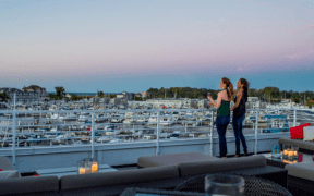 Two women stand next to a railing on the Marina Grand Rooftop Patio Lounge overlooking the harbor with sailing boats.