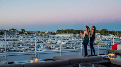 Two women stand next to a railing on the Marina Grand Rooftop Patio Lounge overlooking the harbor with sailing boats.