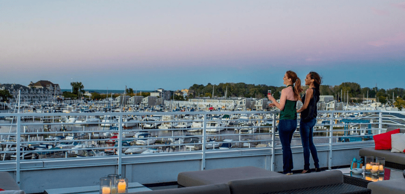 Two women stand next to a railing on the Marina Grand Rooftop Patio Lounge overlooking the harbor with sailing boats.