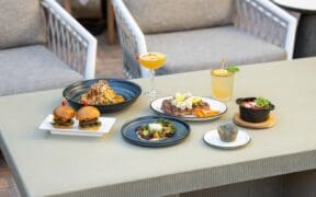 A table with various dishes; burgers, muffin, pasta and cocktails are on a patio table with two chairs at The Wigwam Arizona.
