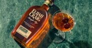 A bottle of Elijah Craig whiskey laying flat on a table next to a full martini glass full of whiskey with a double cherry garnish.