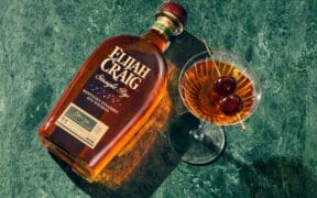 A bottle of Elijah Craig whiskey laying flat on a table next to a full martini glass full of whiskey with a double cherry garnish.