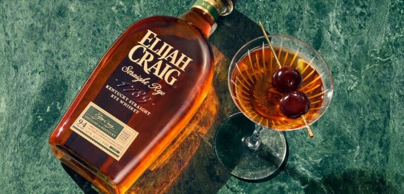 A bottle of Elijah Craig whiskey laying flat on a table next to a full martini glass full of whiskey with a double cherry garnish.