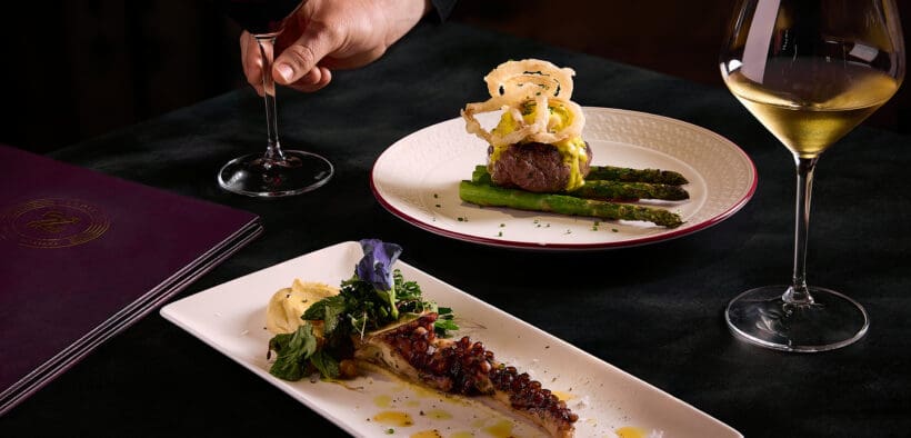 A person holds the stem of a red wine glass. Octopus and steak entrees are on the table with a glass of white wine.