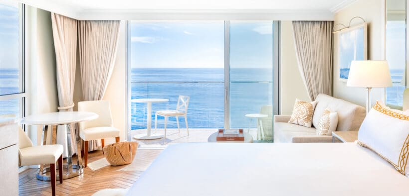 A king-size bed, a couch, a table and two chairs with glass-front windows at Surf & Sand, Laguna Beach looking out to Laguna Beach.