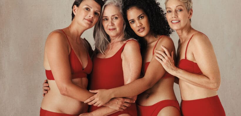 Four midlife-aged women of different ethnicities wearing bras and underwear hug.