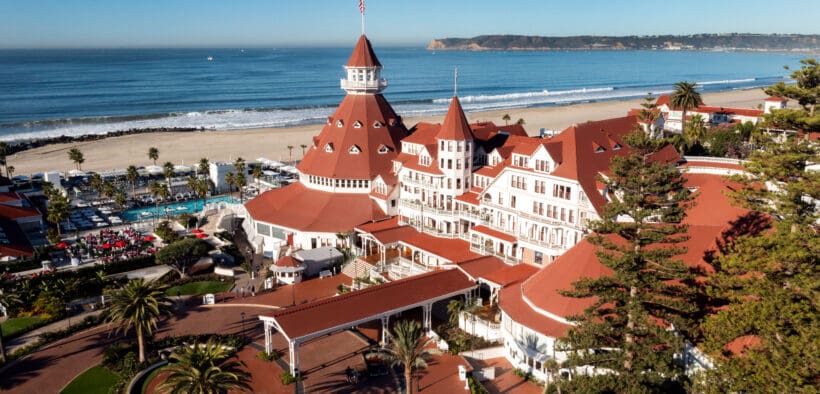 An aerial view of Hotel Del Coronado with its turrets and main drive and pool area next to the Pacific Ocean.