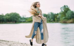 Young blonde woman runs along coastline of sand beach near lake with bouquet of dry flowers in her hands.
