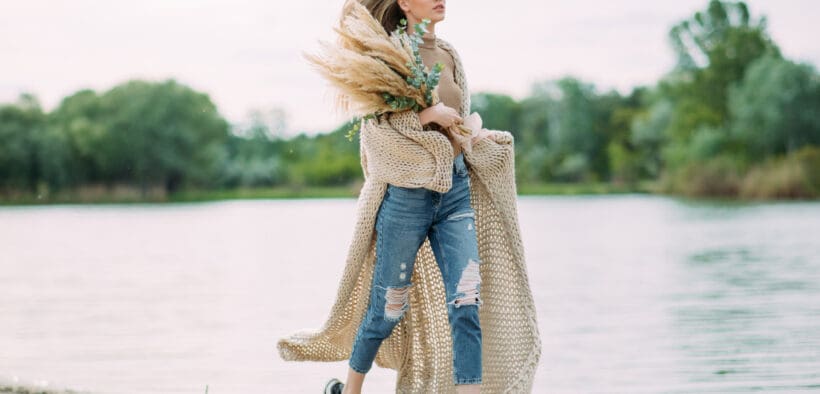 Young blonde woman runs along coastline of sand beach near lake with bouquet of dry flowers in her hands.