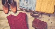A pair of brown boots a red cable sweater, a pair of jeans and yellow cable sweater in a suitcase and a fall fashion purse.