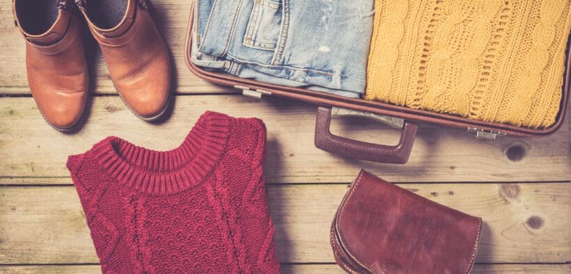 A pair of brown boots a red cable sweater, a pair of jeans and yellow cable sweater in a suitcase and a fall fashion purse.