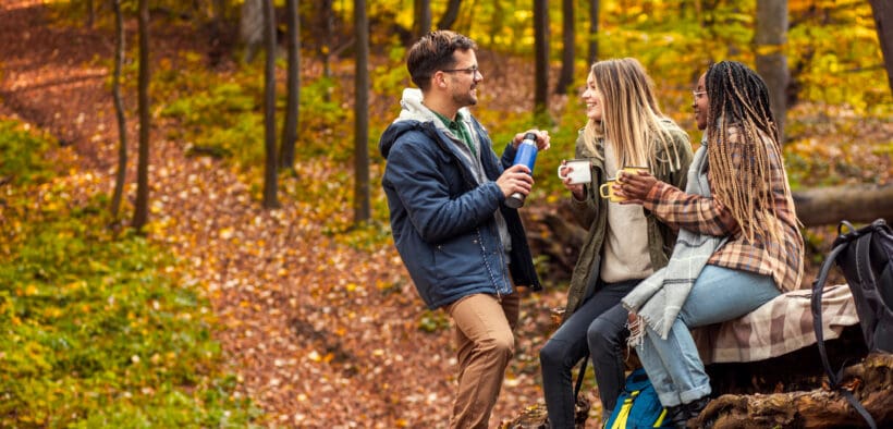 Three friends resting after hiking in forest, sitting on a collapsed trunk and drinking tea.