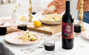 Bricoleur Vineyards 2022 La Rumeur Red Blend bottle on a table next to glasses half full of the wine and two people with dinner plates in front of them.