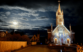 A lit up church near a fenced neighborhood at night, with dark clouds and the moon in Virginia City.