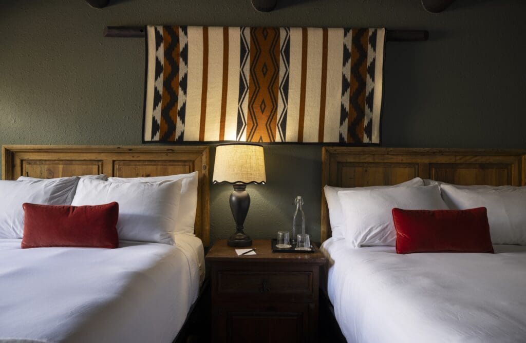 Two beds, each with a throw pillow separated by a nightstand with lamp and native blanket wall hanging in a guestroom at Hotel Glorieta. 