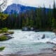 The Canadian Rockies with a mountain peek behind tall pine trees and rushing spring water.