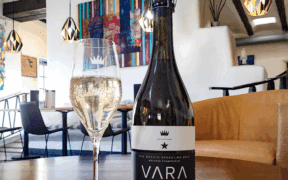A bottle of Vara wine next to a glass of sparkling wine with the VARA Winery tasting area in the background.