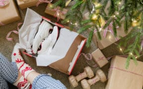 A person with pajama pants and Christmas socks opens a pressent with shoes inside and dumbbells nearby under a Christmas tree.