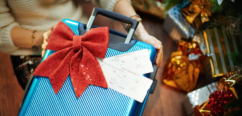 A woman receives a suitcase with two plane tickets as a gift from under the Christmas tree.