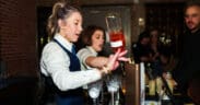 A bartender pours a bottle of liquor into glass with another bartender in the background making a drink at Arizona Cocktail Weekend.