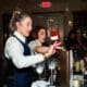 A bartender pours a bottle of liquor into glass with another bartender in the background making a drink at Arizona Cocktail Weekend.
