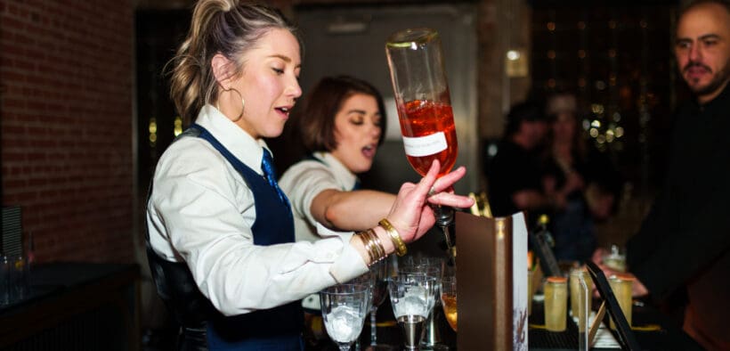 A bartender pours a bottle of liquor into glass with another bartender in the background making a drink at Arizona Cocktail Weekend.