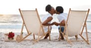 Happy Black couple spending a day at the sea together, kissing from their beach chairs.