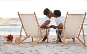 Happy Black couple spending a day at the sea together, kissing from their beach chairs.