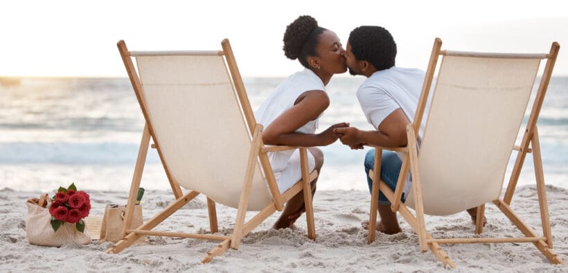 Happy Black couple spending a day at the sea together, kissing from their beach chairs.