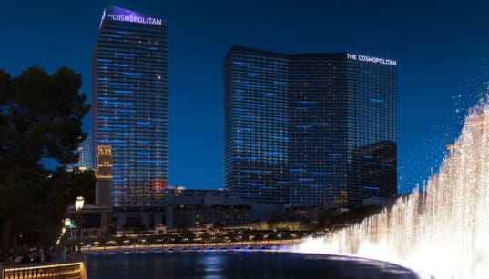 Exterior of The Cosmopolitan of Las Vegas at night shows two buildings with 40 floors each in front a lake with fountains.