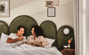 Couple laughing together in bed, holding coffee mugs and a book on a Valentine’s Day date night in downtown Denver.
