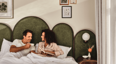 Couple laughing together in bed, holding coffee mugs and a book on a Valentine’s Day date night in downtown Denver.