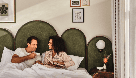Couple laughing together in bed, holding coffee mugs and a book on a Valentine’s Day date night in downtown Denver.