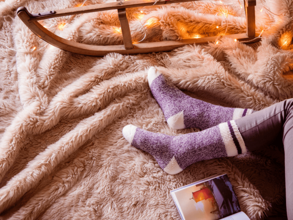 A pair of Heat Holders socks on a woman's feet, with a pink plush rug, rocking chair bottom and magazine on the floor.