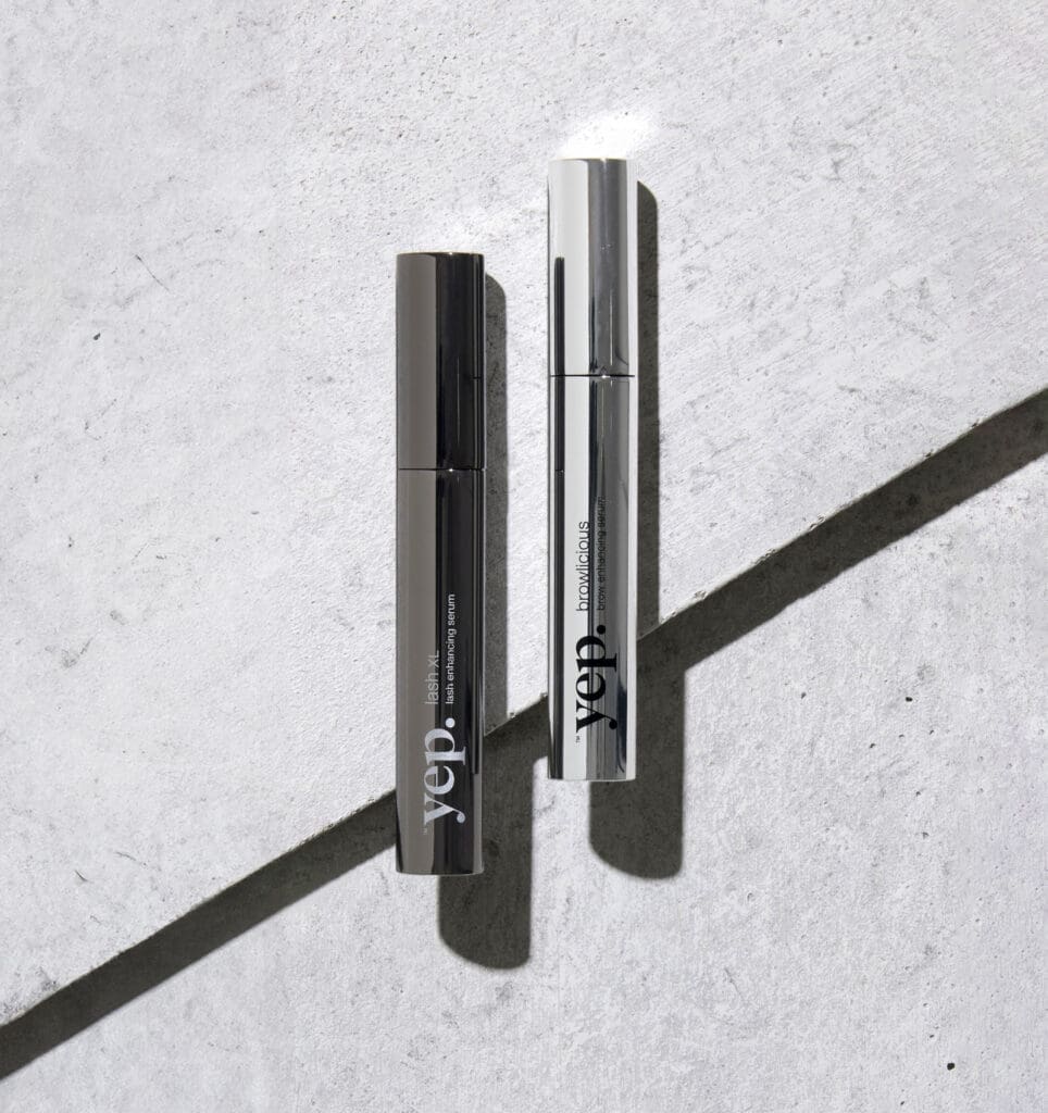 Yep Lash XL eyelash serum and Browlicious eyebrow serum displayed in sleek silver tubes against a textured neutral background.