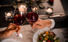Couple clinking glasses of red wine over a candlelit dinner table with salad plates and soft romantic lighting in the background.