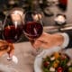 Couple clinking glasses of red wine over a candlelit dinner table with salad plates and soft romantic lighting in the background.