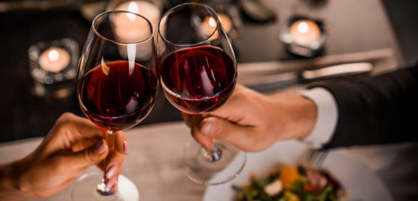 Couple clinking glasses of red wine over a candlelit dinner table with salad plates and soft romantic lighting in the background.