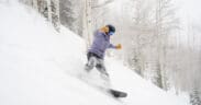Snowboarder carving through deep fresh powder on a tree-lined run at Aspen Snowmass during a winter storm.