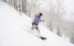 Snowboarder carving through deep fresh powder on a tree-lined run at Aspen Snowmass during a winter storm.