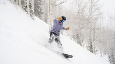 Snowboarder carving through deep fresh powder on a tree-lined run at Aspen Snowmass during a winter storm.