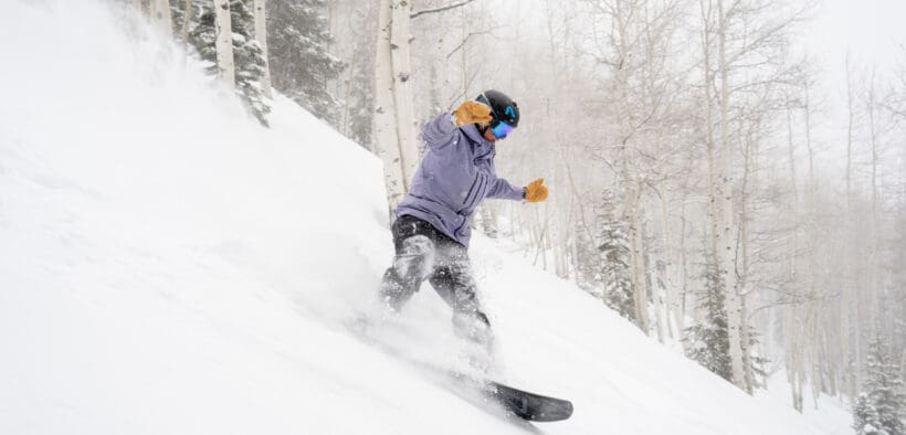 Snowboarder carving through deep fresh powder on a tree-lined run at Aspen Snowmass during a winter storm.
