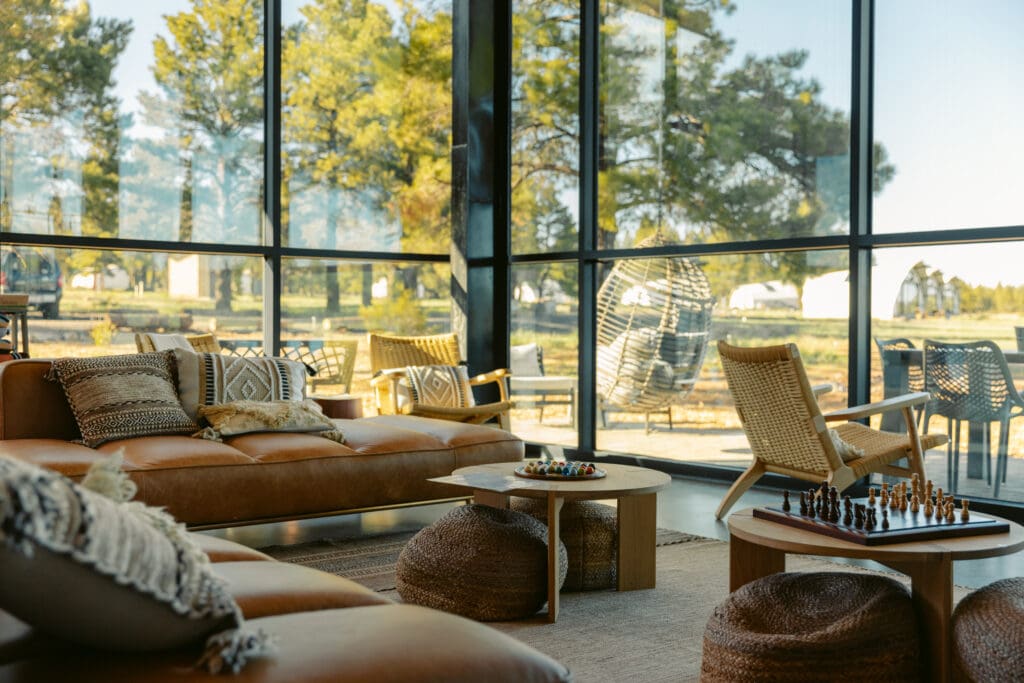 Modern lounge area inside Backland Luxury Nature Resort featuring leather seating, woven chairs and floor-to-ceiling glass walls overlooking forested landscape in Northern Arizona.