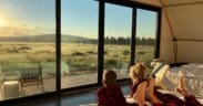 Two children wearing red plaid pajamas lie on a bed inside a luxury glamping tent at Backland Luxury Nature Resort, looking out through large glass doors at a golden sunrise over open meadow and forest in Northern Arizona.