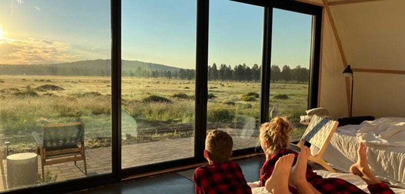 Two children wearing red plaid pajamas lie on a bed inside a luxury glamping tent at Backland Luxury Nature Resort, looking out through large glass doors at a golden sunrise over open meadow and forest in Northern Arizona.