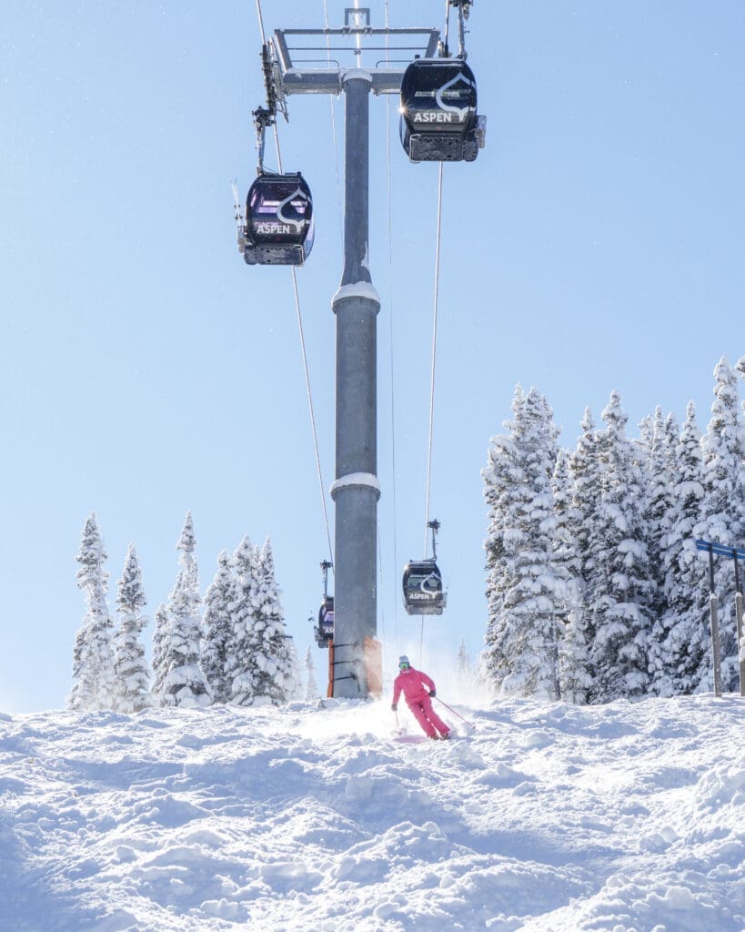 Skier in bright pink gear carving through fresh snow beneath Aspen gondola cabins on a sunny winter day surrounded by snow-covered trees.